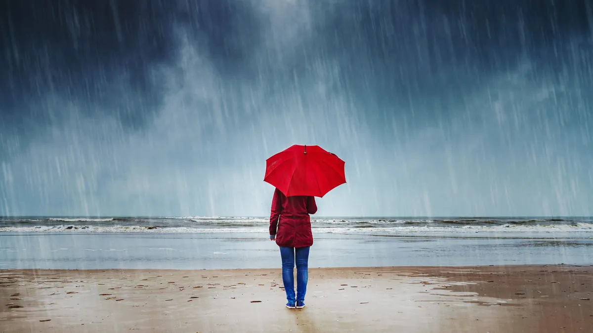 Eine Urlauberin steht mit Regenschirm am Strand (Symbolbild): Gegen schlechtes Wetter kann man sich mittlerweile versichern.